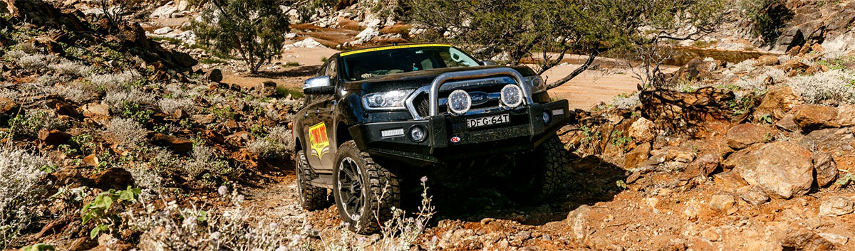 A Ford Ranger with lift kit drives through rocky terrain.